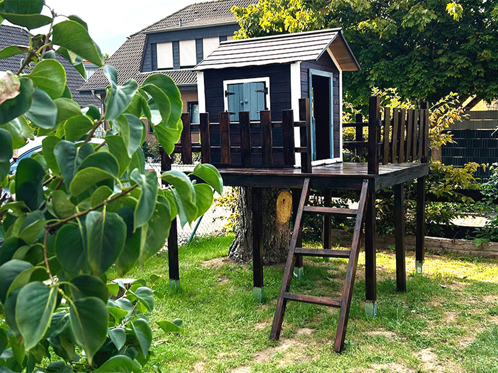 Selbstgebautes Baumhaus aus dunklem Holz mit Terrasse und kleiner Leiter, auf Stelzen rund um einen Baum im Garten errichtet.