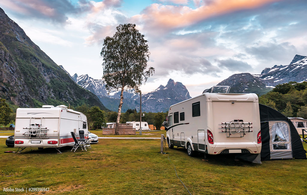 Ein Campingplatz mit Wohnwagen und Wohnmobilen vor einer malerischen Berglandschaft mit schneebedeckten Gipfeln und einem bunten Abendhimmel.