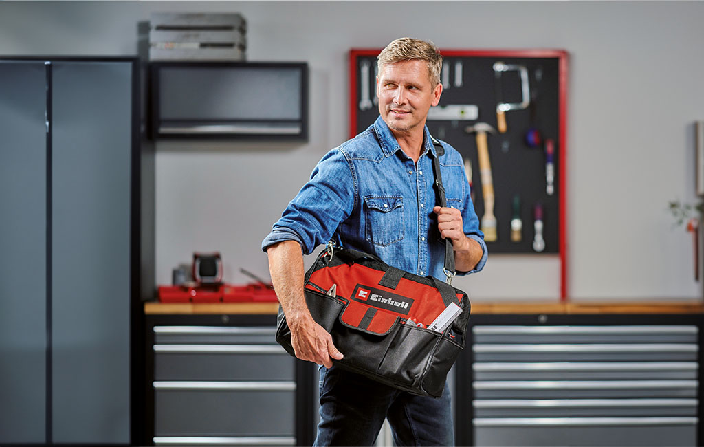 Ein Mann in einem blauen Jeanshemd trägt eine Einhell-Werkzeugtasche in einer Werkstatt mit Werkzeugen im Hintergrund.