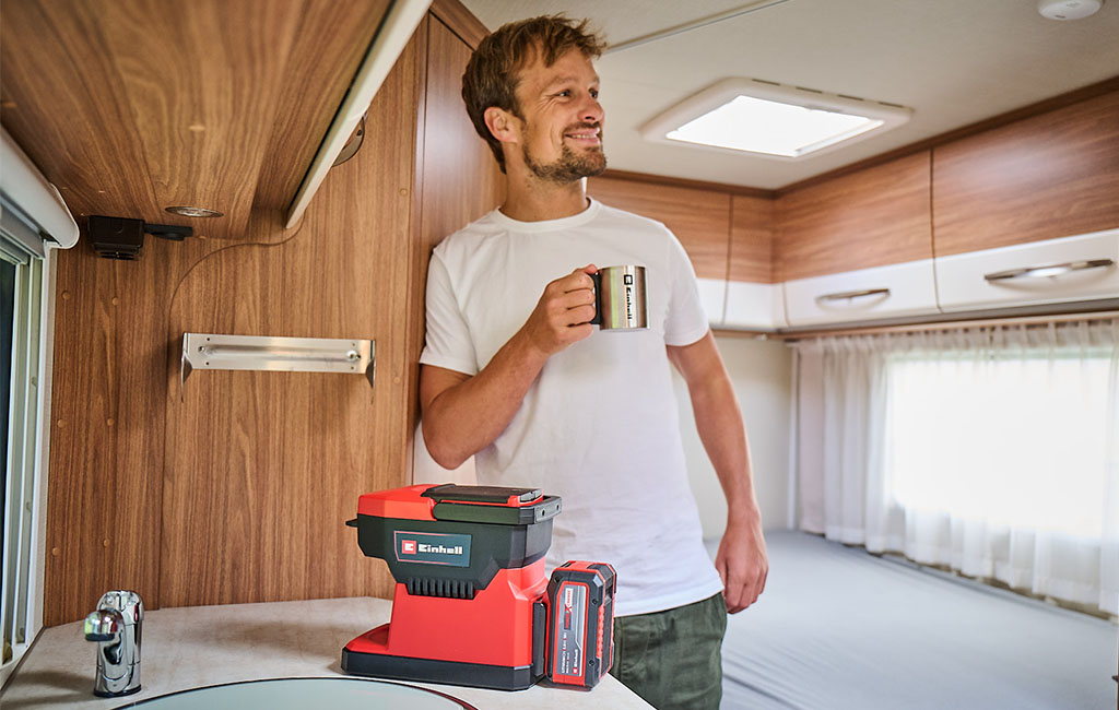 Ein Mann steht in einem Wohnmobil, hält eine Tasse und lächelt, während die Einhell Akku-Kaffeemaschine auf der Küchentheke vor ihm steht.