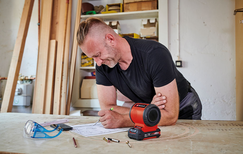 Ein Mann arbeitet in einer Werkstatt, beugt sich über einen Tisch und schreibt auf ein Blatt Papier, während der Einhell Akku-Lautsprecher und eine Schutzbrille neben ihm liegen.