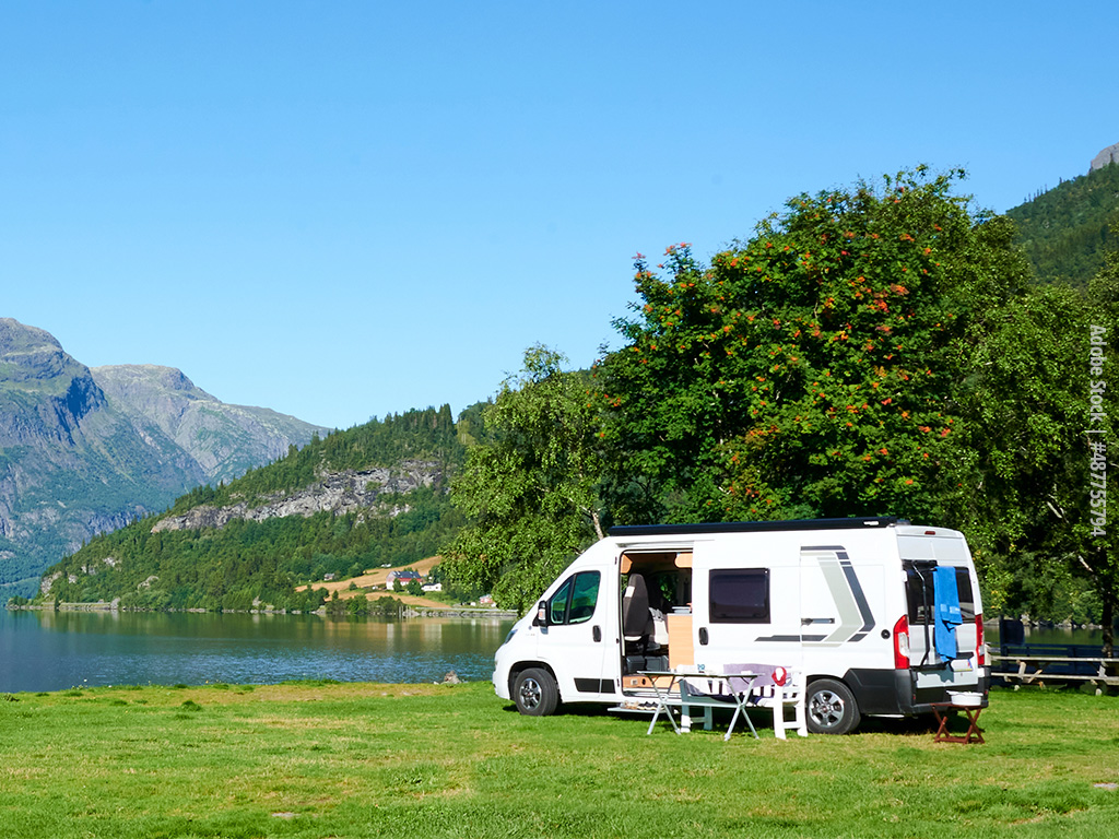 A motorhome is parked on a campsite on the shore of a lake, surrounded by green trees and high mountains under a clear blue sky.