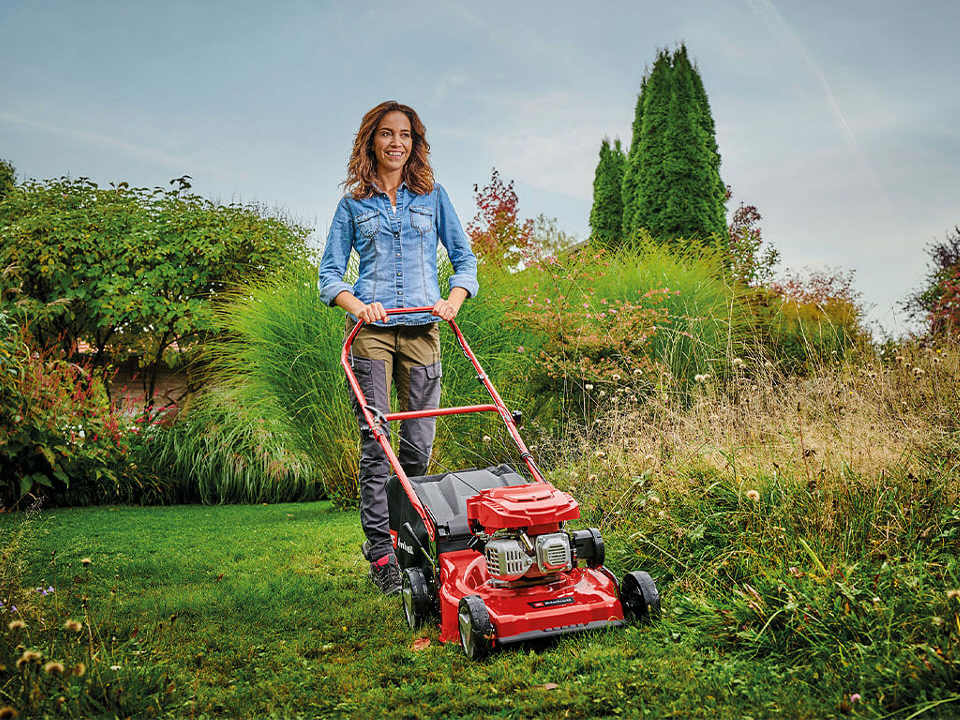 Eine Frau mäht sehr hohes Gras mit einem kräftigen Benzin-Rasenmäher von Einhell.