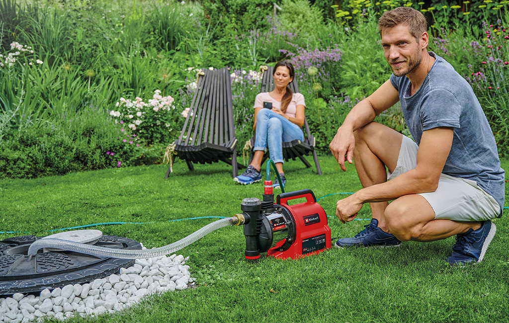 A man kneels next to a garden pump while a woman sits in the background.