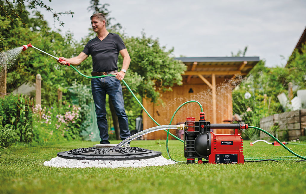 A man sprays water in a garden with an automatic garden pump in the foreground.