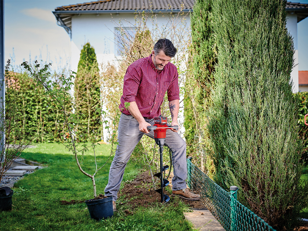 A man using a cordless earth auger to drill a hole into the soil of his garden.