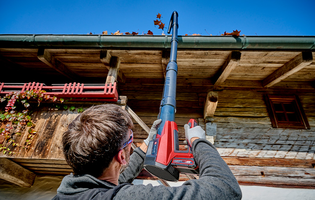 A man uses an Einhell cordless leaf blower with an extension to remove leaves from a gutter from the ground.