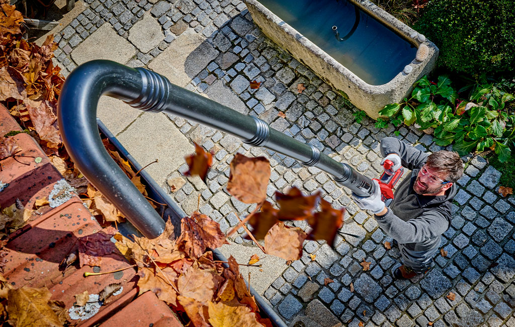 A man uses a leaf blower with an extension to blow leaves out of a gutter while standing on the ground and looking up.