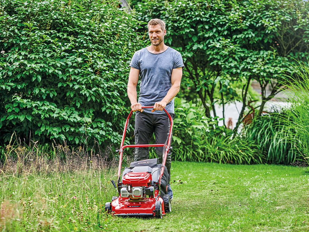 A man pushes his Einhell petrol lawnmower through the garden.