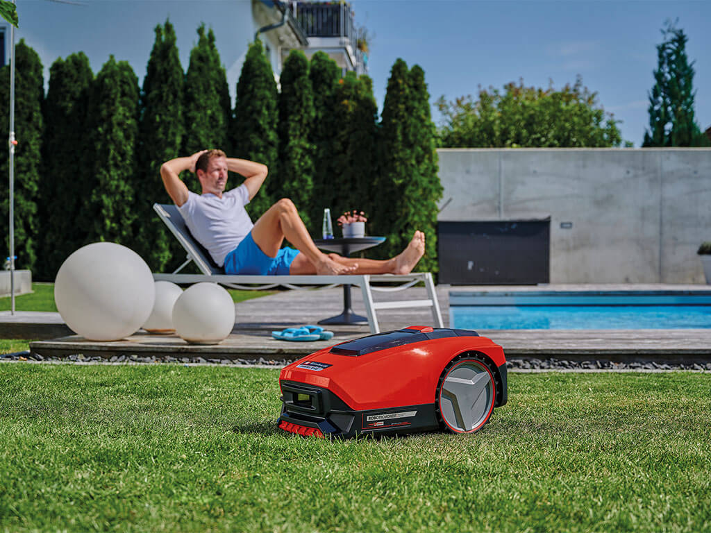 A man lies on a sun chair in the garden, in the foreground the Einhell FREELEXO robotic mower is mowing the lawn.