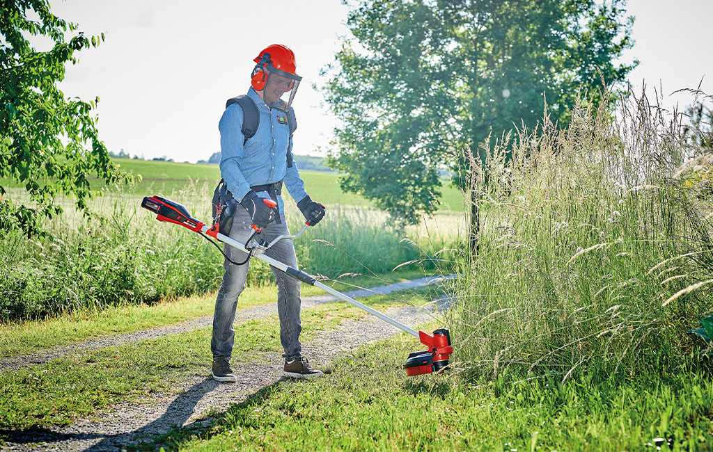 A man with a safety helmet and visor is working with an Einhell cordless brush cutter, cutting tall grass by a field path.