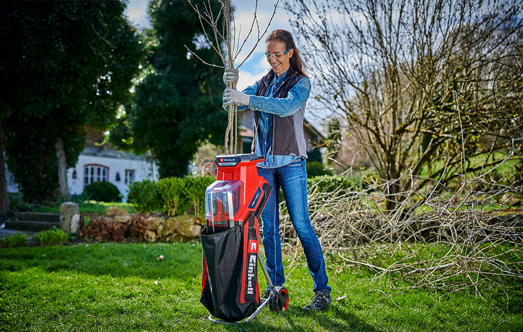 Eine Frau zerkleinert Äste mit einem Einhell Akku-Häcksler in einem Garten, umgeben von Laubhaufen und kahlen Bäumen.