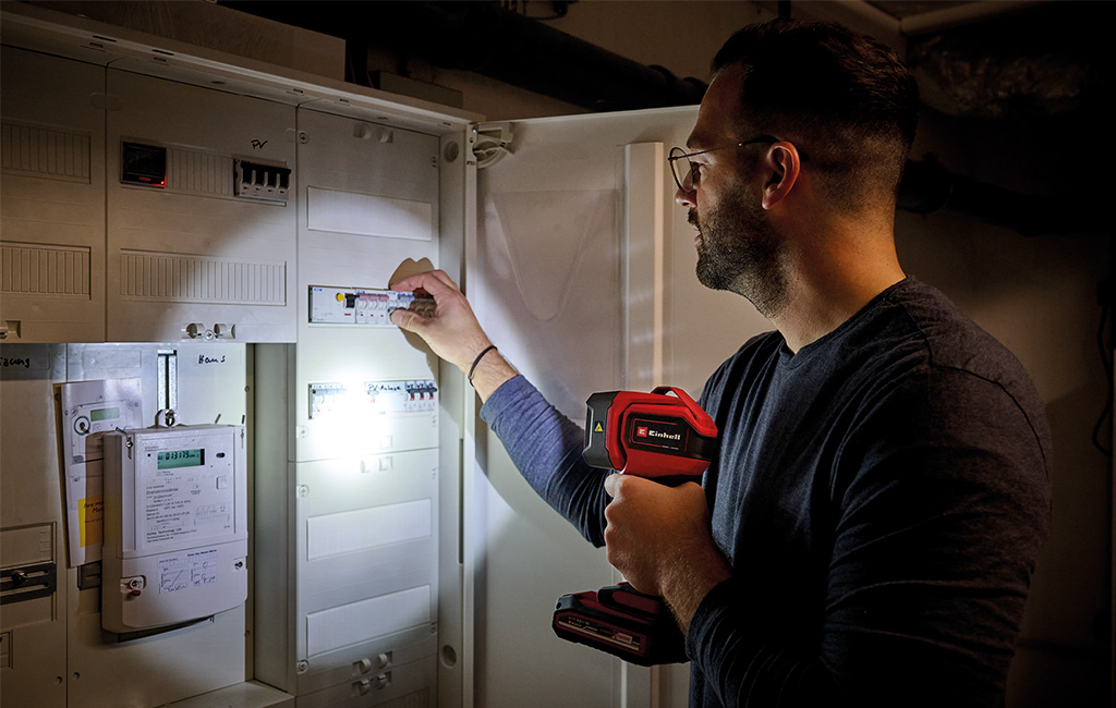 Ein Mann beleuchtet einen Stromkasten mit einer Einhell Akku-Lampe, während er eine Sicherung überprüft.