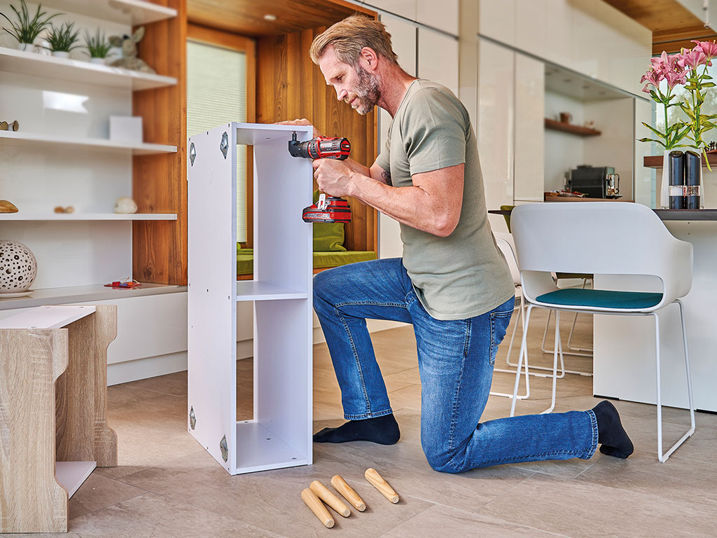 A man assembles a shelf in a living room using an Einhell cordless drill.