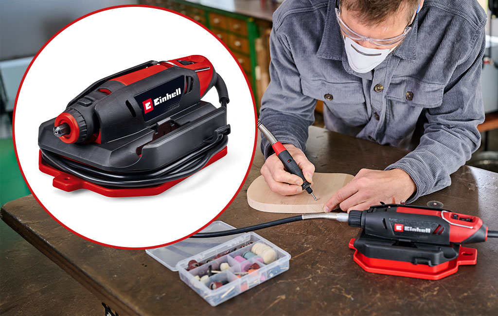 A man wears safety glasses and a mask while working on wood with a red engraving tool.