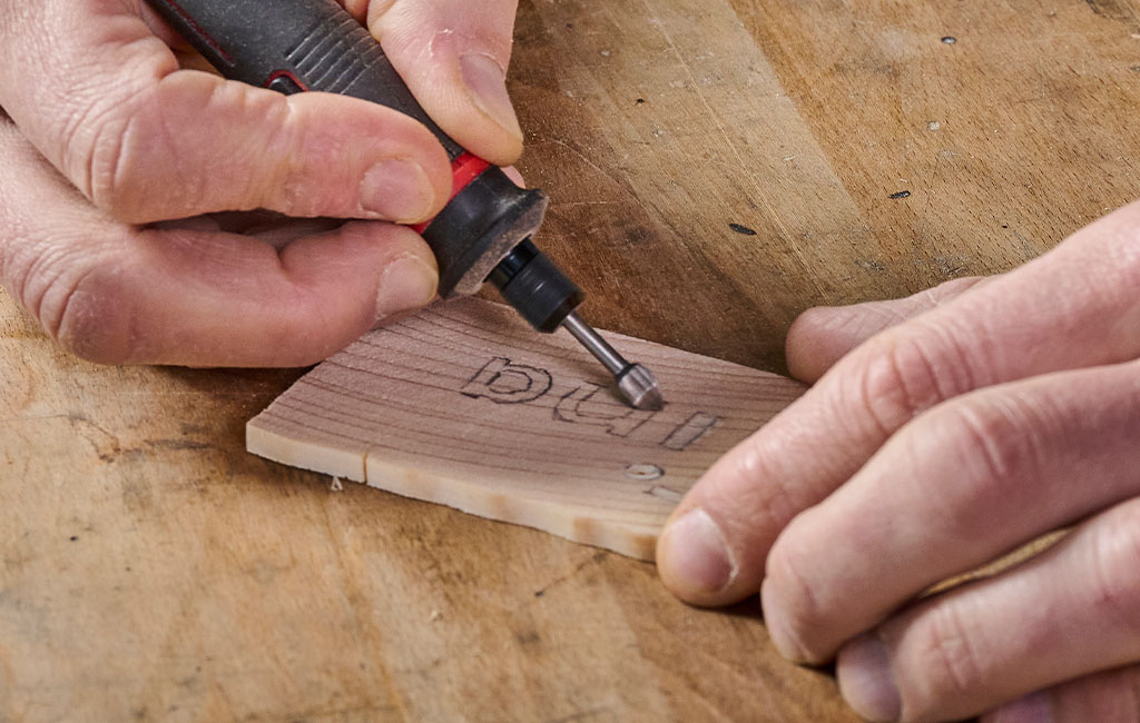 Close-up of hands engraving letters into a piece of wood using a red Einhell engraving tool.