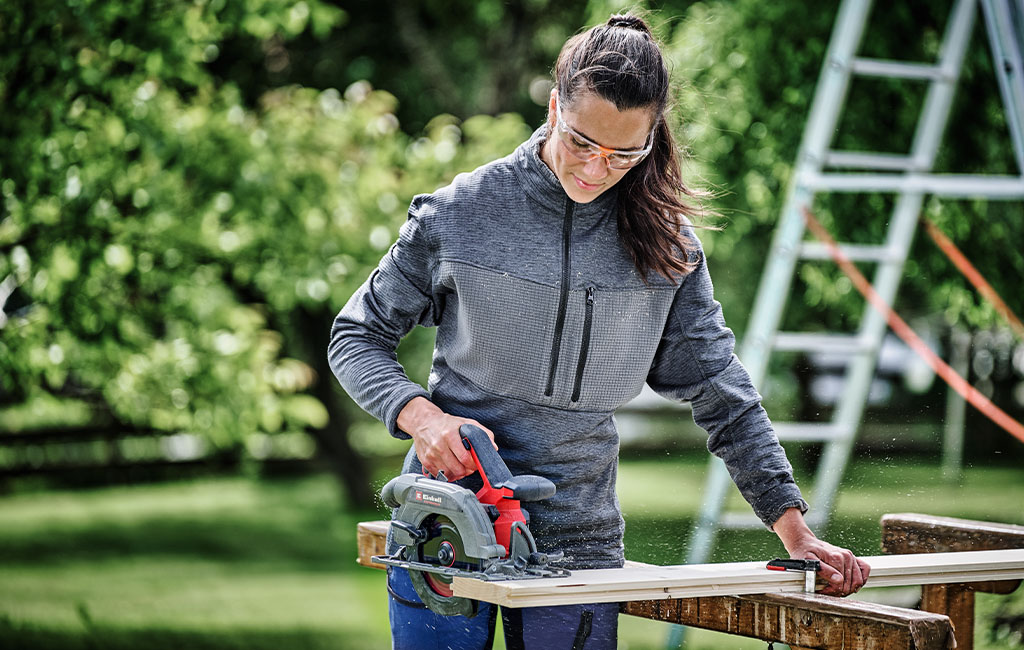 Eine Frau mit Schutzbrille benutzt eine Handkreissäge, um Holz draußen zu schneiden.