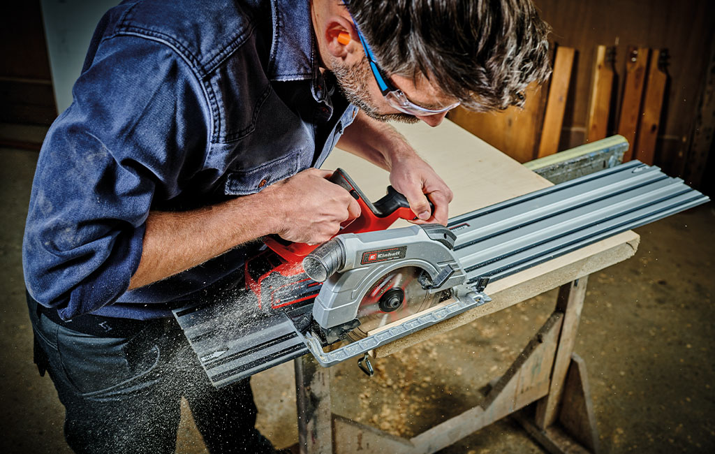 A man wearing safety glasses is cutting wood with a handheld circular saw on a guide rail.