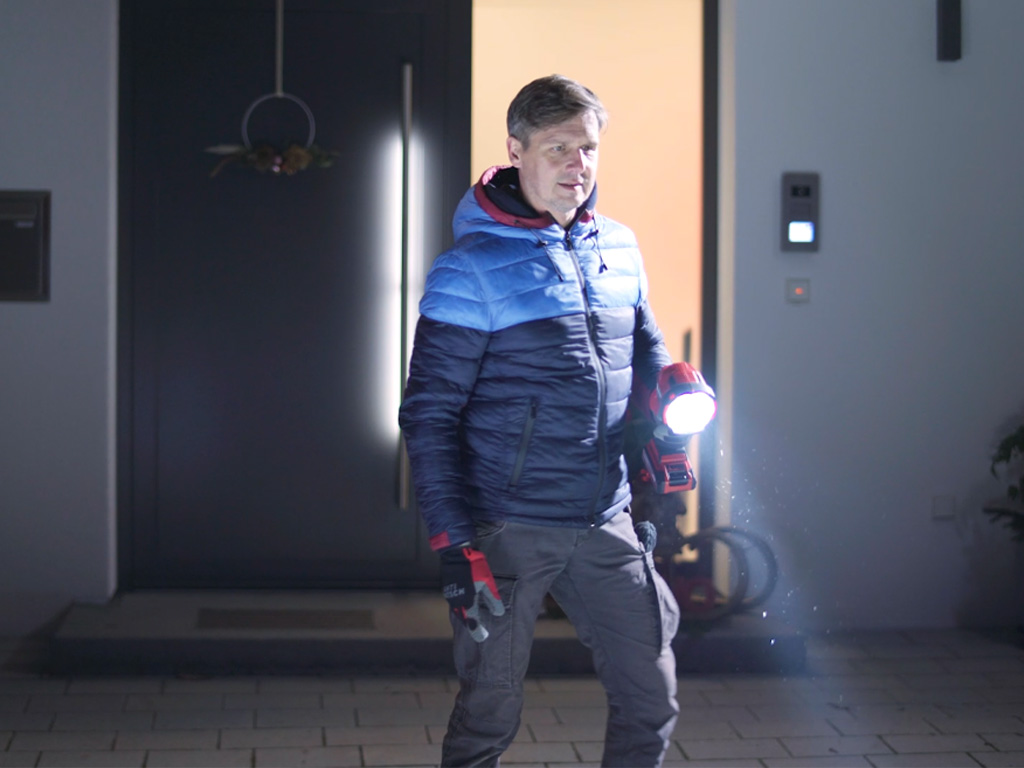 A man carries a work light in the dark in front of a house.