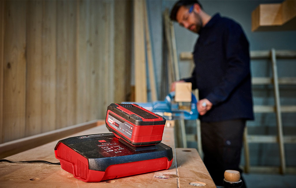 in the front: a Einhell battery charger with a battery | in the back: a man working with a cordless tool