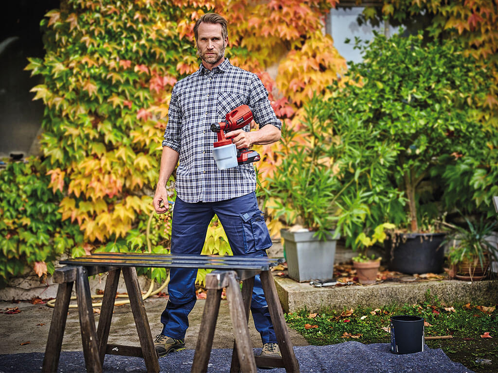 A man stands in front of painted wooden slats and holds a cordless paint sprayer from Einhell in his hand.