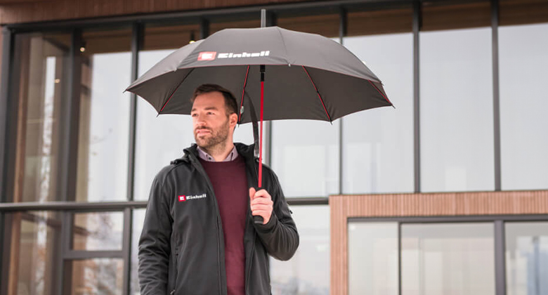 Ein Mann mit Einhell Jacke hält einen Einhell Regenschirm vor einem modernen Gebäude.