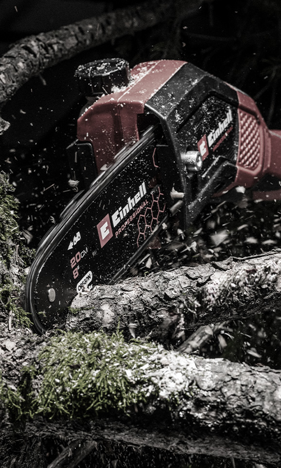 A red Einhell cordless chainsaw cuts through a branch with sawdust flying through the air.