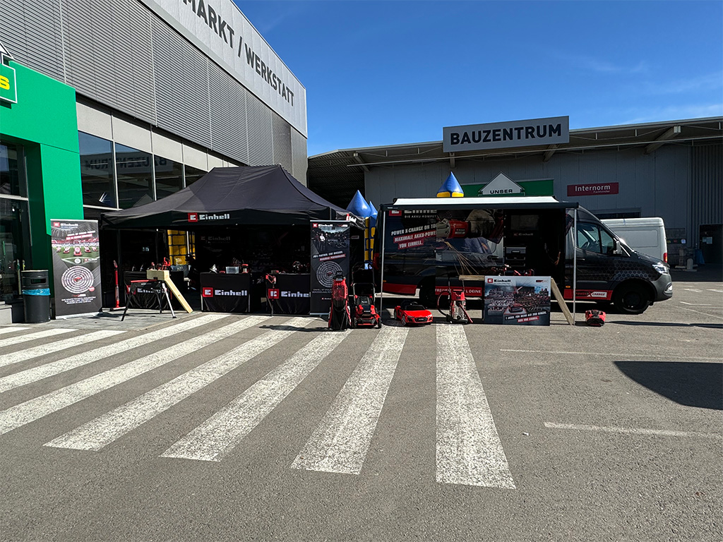 Einhell tent showcasing tools and promotional materials, set up in front of a "Bauzentrum" hardware store.