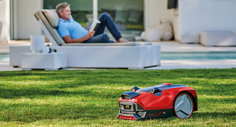 A man lying on a lounger in the garden, a robotic mower cutting the lawn in front of him.